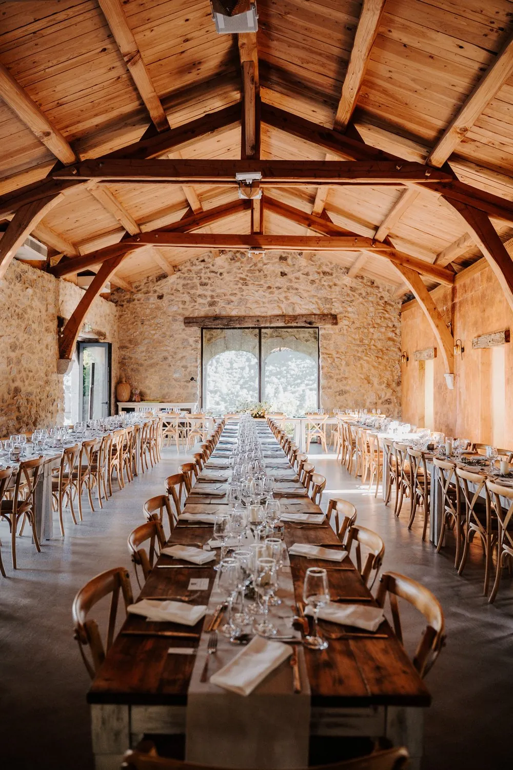 Une longue tablée dressée pour un dîner de mariage, dans une salle en pierre et charpente bois