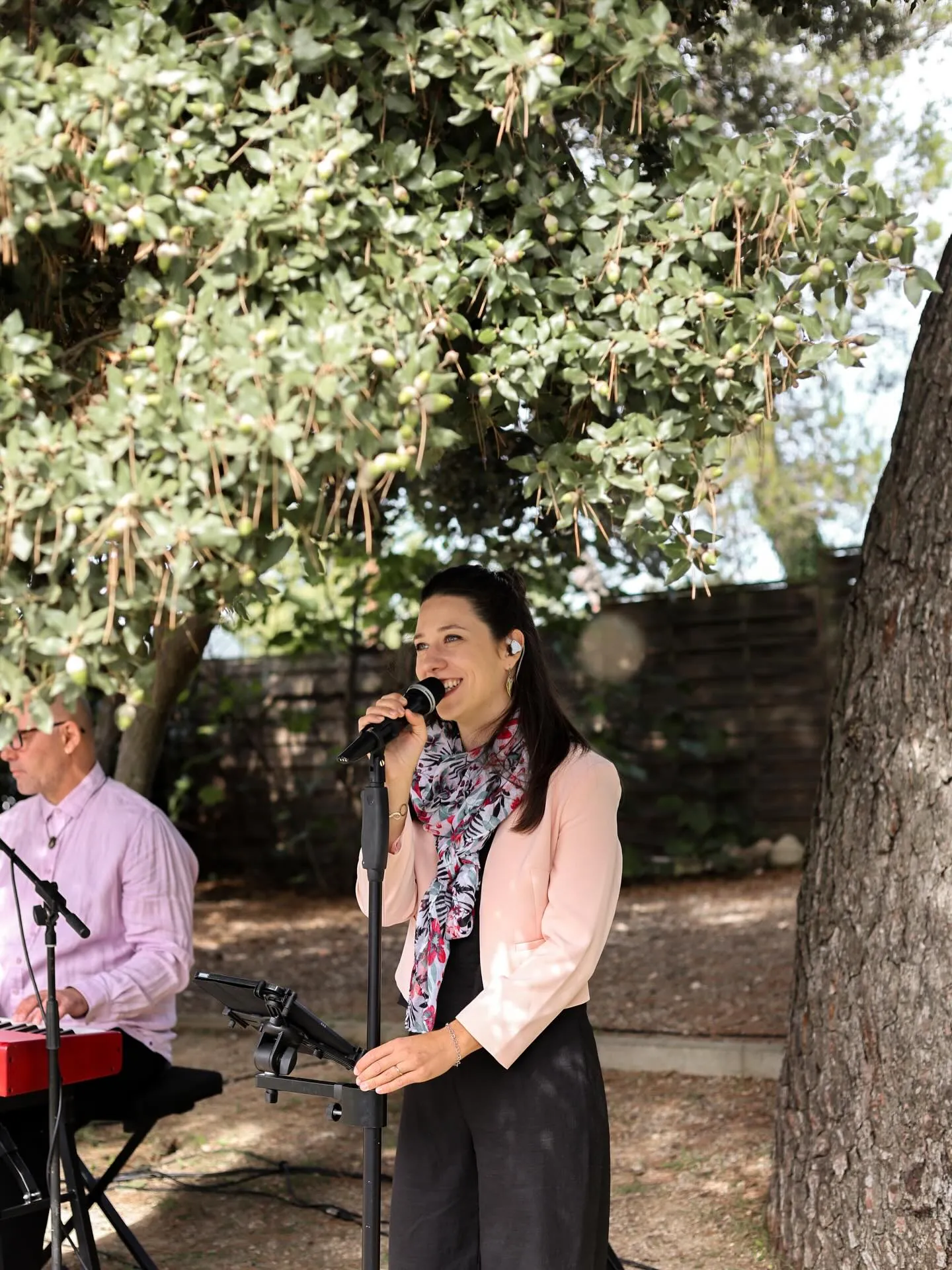 Mathilde chante au micro sous un grand arbre lors d'un cocktail de mariage en extérieur, Stéphane au piano en arrière-plan